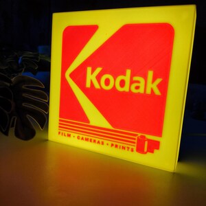May include: A yellow and red Kodak sign. The sign has a yellow border and a red central design with the Kodak logo in white. The text "FILM &bull; CAMERAS &bull; PRINTS" is below the logo. The sign is illuminated.