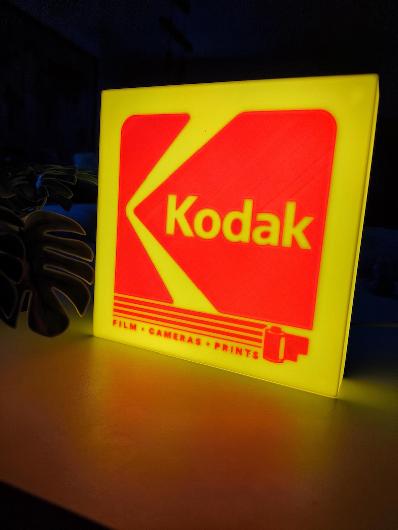 May include: A yellow and red Kodak sign. The sign has a yellow border and a red central design with the Kodak logo in white. The text "FILM &bull; CAMERAS &bull; PRINTS" is below the logo. The sign is illuminated.