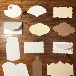 Op de afbeelding: Een set van 12 blanco papieren stansen in verschillende vormen en maten, waaronder een palet, een waaier, een pot, een fles, een boek en verschillende frames. De stansen zijn gemaakt van wit, bruin en crèmekleurig papier.
