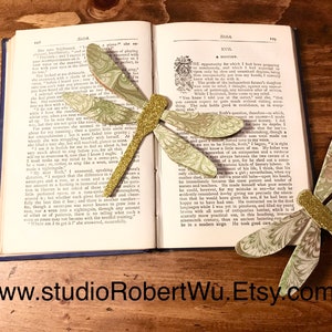 Pode incluir: Dois marcadores de página de libélula verde e dourado com asas de papel marmorizado. Os marcadores de página estão colocados em um livro aberto.