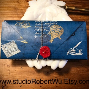 May include: A navy blue gift box with gold and white vintage-style illustrations, including a hot air balloon and typewriter. The box is sealed with a red wax seal and tied with twine. The website address www.studioRobertWu.Etsy.com is at the bottom.