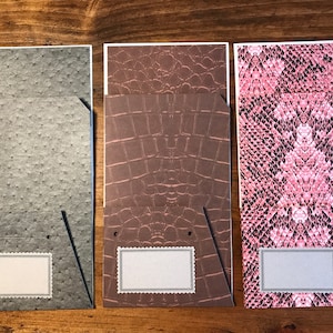 May include: Three decorative folders with different patterns. The folder on the left has a gray ostrich skin pattern, the middle folder has a brown crocodile skin pattern, and the folder on the right has a pink snake skin pattern. Each folder has a white label with a black border.