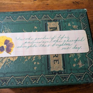 Puede incluir: Un libro antiguo con una cubierta verde azulado y detalles dorados. Un pensamiento amarillo y morado prensado está adherido a una etiqueta blanca con texto manuscrito: "En el jardín de la vida, los pensamientos son los pensamientos alegres que iluminan nuestro día."