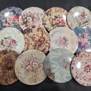 May include: A collection of twelve round coasters with floral designs. The coasters feature various color schemes, including blue, cream, and brown, with detailed rose and floral patterns. The coasters are likely made of a durable material.