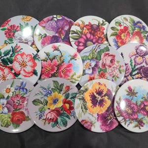 May include: A collection of twelve round coasters featuring cross-stitch floral designs. Each coaster showcases a unique arrangement of colorful flowers, including roses, pansies, and irises, in shades of red, pink, purple, and yellow, set against a light gray background.