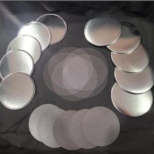 May include: An overhead shot displays a collection of silver metal lids, clear circular sheets, and gray circular sheets. The lids are arranged in groups, while the sheets are layered in the center and bottom. The background is a dark, textured surface.