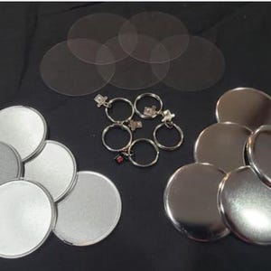 May include: An assortment of crafting supplies, including silver-colored metal button blanks, clear plastic circles, and keychains with charms. The items are arranged on a black surface, suitable for creating custom buttons or accessories.