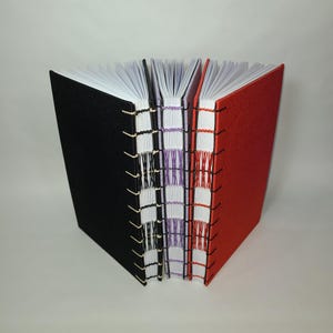 May include: A collection of handmade journals with exposed binding. The covers are black, purple, and red. The pages are white, and the binding thread matches the cover colors. The journals are fanned out, showcasing the detailed stitching.
