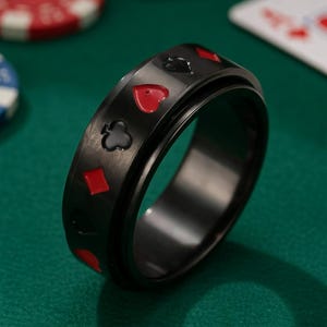 Può includere: Anello nero con simboli di carte da gioco rossi e neri. L'anello presenta una fascia rotante con simboli di picche, cuori, quadri e fiori. Su una superficie verde, con fiches da poker e una carta da gioco sullo sfondo.
