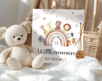 May include: A cream-colored knitted teddy bear sits next to a children's book with a rainbow and animal illustrations. The book has the German text "Willkommen auf der Welt" and a small tag that says "Alles Gute".