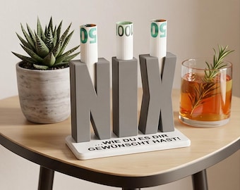 May include: A gray decorative stand featuring the letters N, I, and X, each with a cylindrical opening holding rolled-up currency. The stand is mounted on a white rectangular base with the black text ...WIE DU ES DIR GEWÜNSCHT HAST!