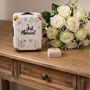 May include: A white miniature suitcase with gold-toned corners, featuring black line art of birds, a car, and the text "Just Married" with hearts. Beside it sits a beige ring box holding two gold bands and a bouquet of white roses.