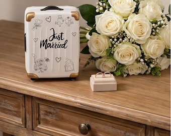 May include: A white miniature suitcase with gold-toned corners, featuring black line art of birds, a car, and the text "Just Married" with hearts. Beside it sits a beige ring box holding two gold bands and a bouquet of white roses.