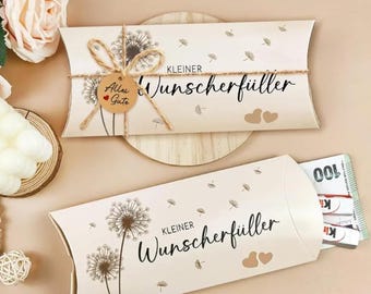 May include: Two beige gift boxes with dandelion illustrations and the text "Kleiner Wunschfüller". One box is tied with twine and a small tag that says "Alles Gute". The other box is open, revealing money.