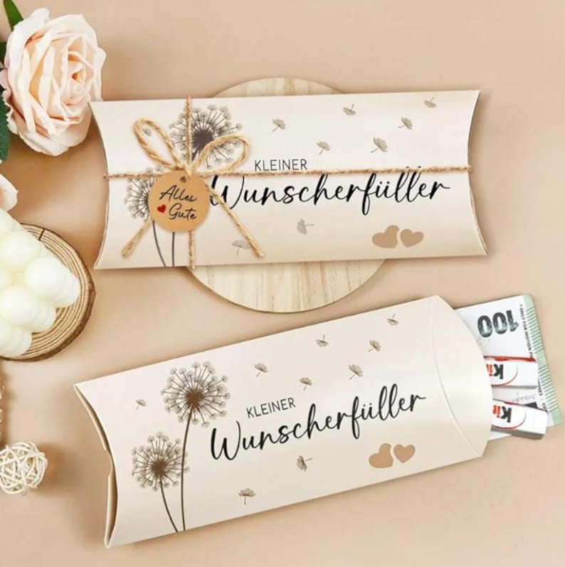 May include: Two beige gift boxes with dandelion illustrations and the text "Kleiner Wunschf&uuml;ller". One box is tied with twine and a small tag that says "Alles Gute". The other box is open, revealing money.