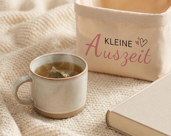 May include: A ceramic mug with tea, a small canvas basket with the text "KLEINE Auszeit" in pink, and a closed book rest on a cream-colored knitted blanket. The mug has a light beige color with a brown base.