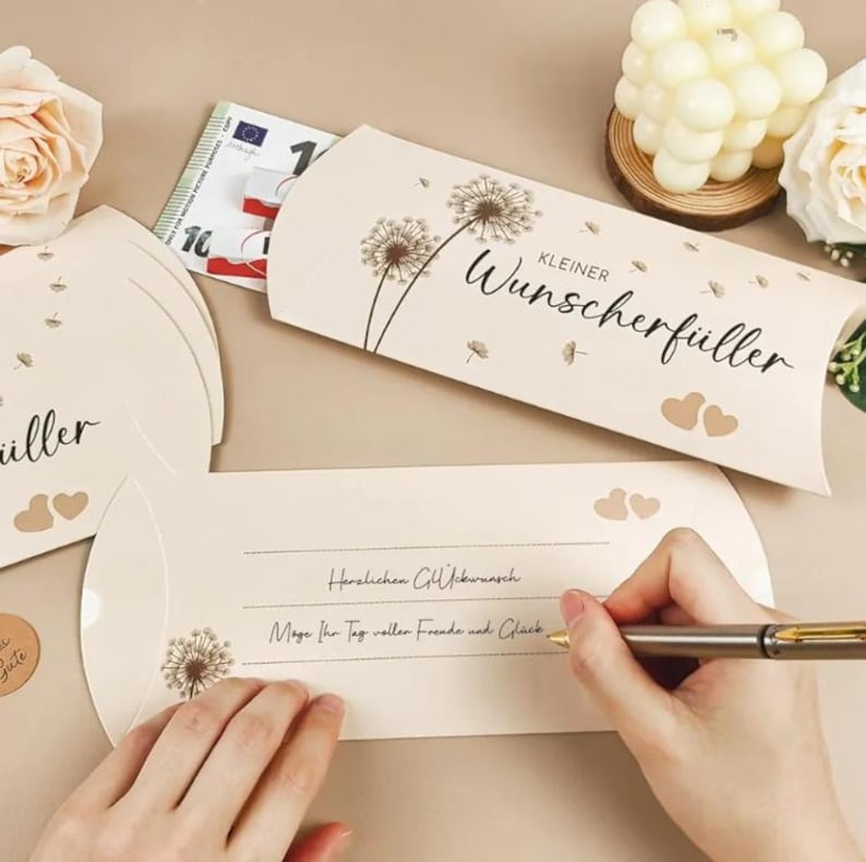 May include: Cream-colored gift card holders with dandelion illustrations and the text "Kleiner Wunscherf&uuml;ller". One holder contains a Euro bill. A person is writing on a card with the text "Herzlichen Gl&uuml;ckwunsch".