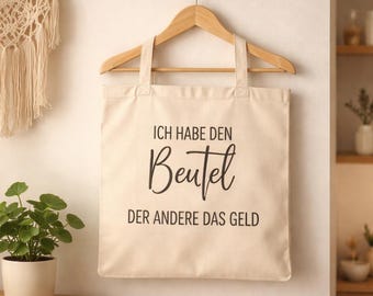 May include: A cream-colored tote bag with black text that reads "ICH HABE DEN Beutel DER ANDERE DAS GELD." The bag hangs on a wooden hanger. A macrame wall hanging and a potted plant are visible in the background.