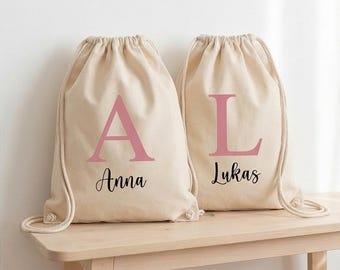 May include: Two beige canvas drawstring backpacks with thick rope straps. The left bag features a large pink letter A and the name Anna in black script. The right bag features a large pink letter L and the name Lukas in black script.