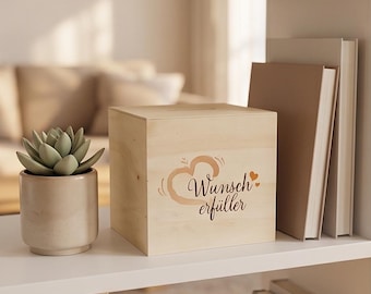 May include: A light-colored wooden box with the words "Wunscherfüller" and a heart design. The box sits on a white shelf next to a small potted succulent and a stack of books. The background is blurred.