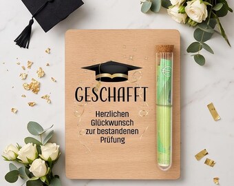 May include: A rectangular wooden plaque featuring a black graduation cap icon and the text "GESCHAFFT Herzlichen Glückwunsch zur bestandenen Prüfung". A clear glass test tube with a cork stopper containing green paper currency is attached.