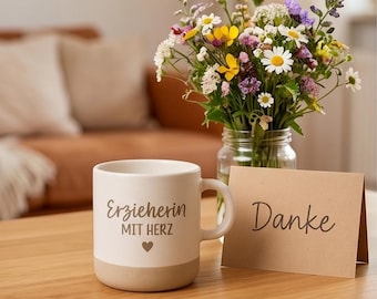 May include: A white and tan ceramic mug with the text "Erzieherin MIT HERZ" and a heart symbol. A small bouquet of colorful wildflowers in a glass jar and a card that says "Danke" are on a wooden table.