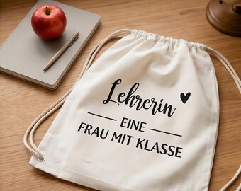 May include: A white drawstring bag with black text that reads "Lehrerin EINE FRAU MIT KLASSE" and a small black heart. A red apple, a gray notebook, and a pen are on a wooden surface.