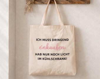 May include: A beige canvas tote bag with the German text "ICH MUSS DRINGEND einkaufen, HAB NUR NOCH LICHT IM KÜHLSCHRANK!" in black and pink lettering. The bag hangs from a wooden rack on a white wall.