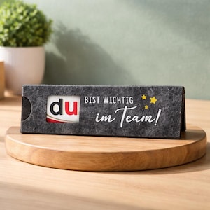 May include: A dark gray felt card holder with the word "du" in red and white, and the text "BIST WICHTIG im Team!" in white. The holder sits on a wooden stand, with a small plant in the background.