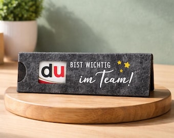 May include: A dark gray felt card holder with the word "du" in red and white, and the text "BIST WICHTIG im Team!" in white. The holder sits on a wooden stand, with a small plant in the background.