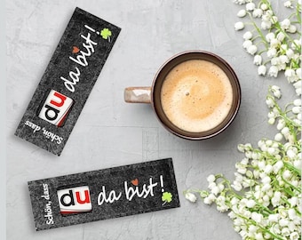 May include: A cup of coffee with a light brown crema sits next to two black rectangular cards with white text that reads "du da bist!" and "Schön, dass". A sprig of white flowers is also present.