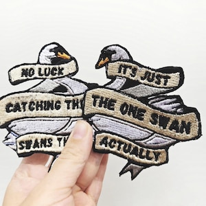 May include: Two embroidered patches featuring white swans with black beaks and eyes. Each patch includes beige banners with black text. The left patch reads NO LUCK CATCHING THE SWANS. The right patch reads IT'S JUST THE ONE SWAN ACTUALLY.