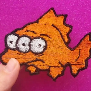 May include: Embroidered orange fish patch with three white eyes and a black outline. The fish has a sad expression. The patch is on a pink background. Suitable for jackets, bags, or other fabric items.