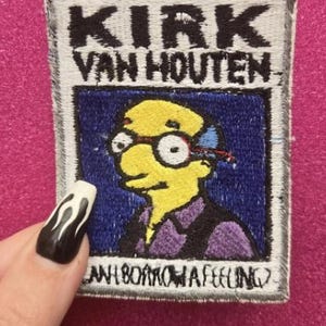 May include: Embroidered patch featuring Kirk Van Houten from The Simpsons. The patch has a white border with black text reading "KIRK VAN HOUTEN." The image inside shows Kirk with yellow skin, glasses, and a purple shirt. The text "CAN I BORROW A FEELING" is at the bottom.