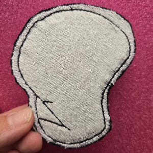 May include: A gray embroidered patch in the shape of a skull with a black outline and a jagged line detail. The patch is held against a pink background.