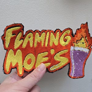 Op de afbeelding: Geborduurde patch met de tekst "Flaming Moe's" in gele draad op een oranje achtergrond. Een paars drankje met vlammen is rechts afgebeeld. De patch heeft een zwarte rand.