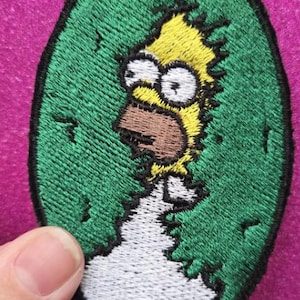 以下が含まれることがあります： 緑色の卵から出てくるホーマー・シンプソンの刺繍パッチ。パッチは黒い縁取りで、ホーマーの黄色い髪、茶色の口、白い目が確認できます。背景は鮮やかなピンク色です。