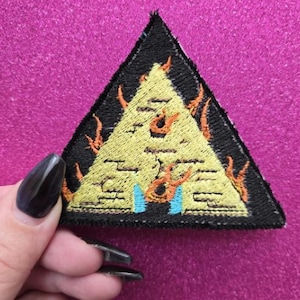 May include: A black, triangular patch featuring a yellow pyramid design with orange flames and blue accents. The patch is embroidered with intricate details and has a black border. The patch is held by a hand with black nail polish.