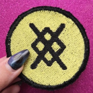 May include: A round embroidered patch with a yellow center and a black border. The patch features a black geometric design. The patch is held against a pink background.