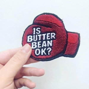 Pode incluir: Um emblema bordado vermelho em forma de luva de boxe com uma borda preta. O texto IS BUTTER BEAN OK? está costurado com linha branca no centro da luva. O emblema apresenta uma superfície texturizada com detalhes de fios visíveis.