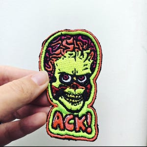 Può includere: Una toppa ricamata raffigurante una testa aliena stilizzata con un grande cervello esposto nei toni del rosso e del rosa. L'alieno ha un volto giallo-verde pallido, occhi scuri e un ampio sorriso. La parola ACK! è ricamata in arancione.