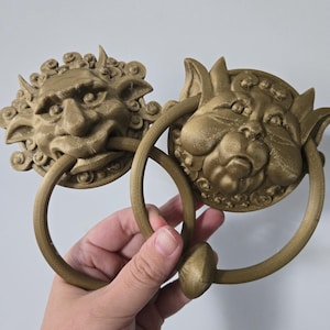 Puede incluir: Dos aldabas de puerta de color bronce, cada una con un diseño de rostro detallado. Una tiene una cara humana y la otra una cara de gato. Ambas tienen un asa circular. Las aldabas se sostienen en una mano sobre un fondo blanco.