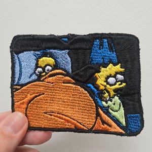 May include: A rectangular embroidered patch featuring a scene with two yellow cartoon characters. One character is lying in a bed under an orange blanket with a blue pillow, while the other character stands to the right in a dark blue setting.