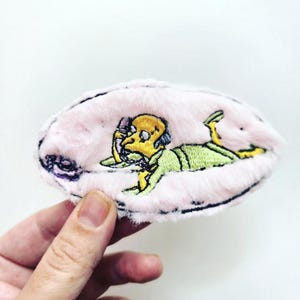 Puede incluir: Un parche ovalado con una textura rosa claro y difusa que presenta un personaje de dibujos animados bordado con piel amarilla, traje verde y cabello oscuro, acostado mientras sostiene un auricular de teléfono contra su oreja.