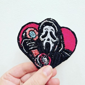 May include: An embroidered patch shaped like a heart featuring a black and white ghost-like figure with a long face and open mouth. The design includes vibrant pink background accents and small circular details in blue and red, all outlined in black.