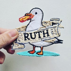May include: An embroidered patch featuring a white seagull with orange feet and beak, holding a hot dog in its mouth. The seagull stands on a teal oval base and is wrapped in a beige banner with the name RUTH embroidered in black capital letters.