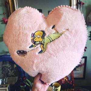 Peut inclure: Un coussin en peluche rose en forme de cœur avec une image de dessin animé d'un personnage jaune en costume vert tenant un téléphone. Le coussin a une bordure de petits pompons roses.