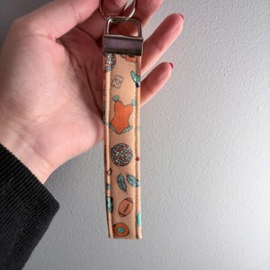 May include: A peach-colored wristlet keychain with a silver-tone metal clasp and key ring. The fabric features a repeating pattern of a disco ball, a football, feathers, and a swimsuit. The wristlet is held by a person.