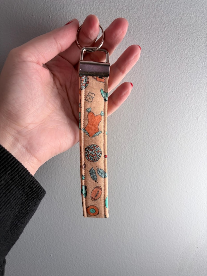 May include: A peach-colored wristlet keychain with a silver-tone metal clasp and key ring. The fabric features a repeating pattern of a disco ball, a football, feathers, and a swimsuit. The wristlet is held by a person.