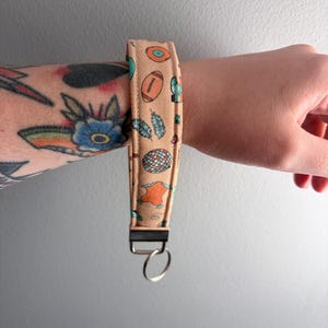 May include: A tan wristlet key fob with a repeating pattern of footballs, feathers, and other small illustrations. The key fob has a black metal clasp and a silver key ring.